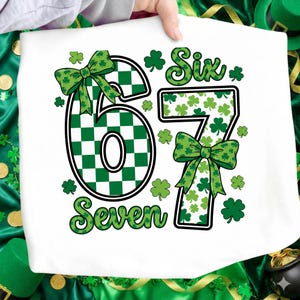 Könnte beinhalten: Weißes T-Shirt mit den Zahlen "6" und "7" in Grün und Weiß. Die Zahl "6" hat ein grün-weißes Karomuster. Das Wort "Six" steht über der "6" und "Seven" unter der "7". Grüne Schleifen und Kleeblätter schmücken das Design.