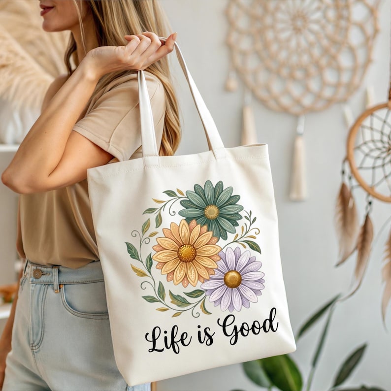 Puede incluir: Bolso tote de color crema con un dise&ntilde;o floral. El dise&ntilde;o presenta tres margaritas en verde, naranja y morado, rodeadas de hojas verdes. Las palabras "Life is Good" est&aacute;n impresas en negro. El bolso tiene asas blancas.