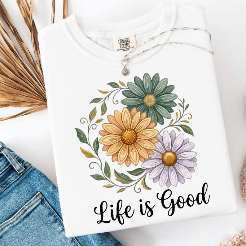 Puede incluir: Sudadera blanca de cuello redondo con un dise&ntilde;o floral con flores amarillas, verdes y moradas. Las palabras "Life is Good" est&aacute;n impresas en negro debajo. Un collar plateado con un colgante de sol descansa sobre la camisa.