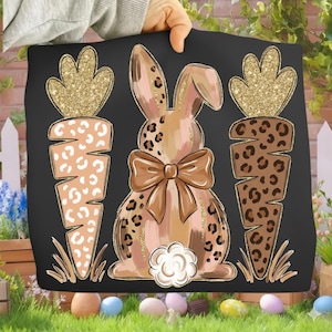 May include: Black square pillow with a bunny and carrot design. The bunny has a bow and a fluffy tail. The carrots have a leopard print design. The bunny and carrots are set against a black background.