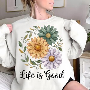 Puede incluir: Sudadera blanca con un dise&ntilde;o floral. Presenta tres margaritas en amarillo, verde azulado y morado, rodeadas de hojas verdes. La frase "Life is Good" est&aacute; impresa en negro debajo.