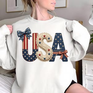 Op de afbeelding: Witte sweatshirt met een patriottisch "USA" ontwerp. De letters zijn versierd met sterren, strepen en strikken in rood, wit en blauw. De "S" is een gebreid ontwerp met rode en blauwe sterren.