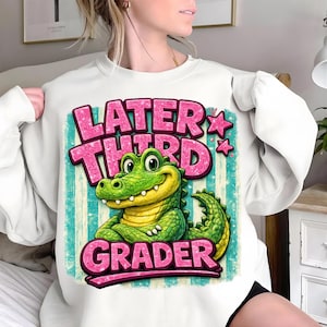 Könnte beinhalten: Weißes Sweatshirt mit einem Cartoon-Alligator und dem Text "Later Third Grader" in Pink. Der Alligator ist grün und gelb, vor einem türkis-weiß gestreiften Hintergrund. Das Design enthält rosa Sterne.