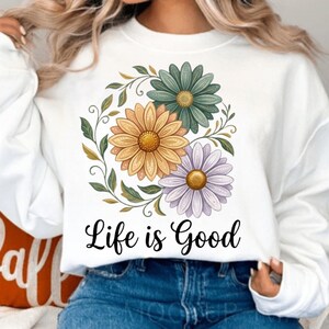 Puede incluir: Sudadera blanca con un dise&ntilde;o floral. Presenta tres margaritas en amarillo, verde y morado, rodeadas de hojas verdes. La frase "Life is Good" est&aacute; escrita en negro debajo de las flores.