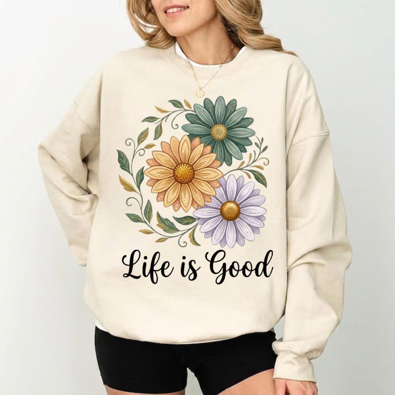 Puede incluir: Sudadera color crema con un dise&ntilde;o floral de margaritas amarillas, turquesas y moradas rodeadas de hojas verdes. Las palabras "Life is Good" est&aacute;n escritas en negro debajo de las flores.