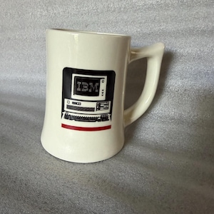 Może przedstawiać: Kremowy kubek ceramiczny z czarnym nadrukiem graficznym przedstawiającym zabytkowy system komputerowy IBM, w tym monitor z logo IBM, jednostkę centralną i klawiaturę, podkreślony cienką poziomą czerwoną linią pod komputerem.