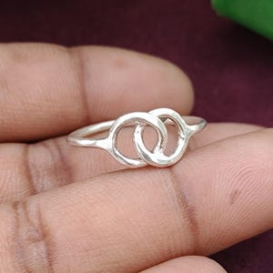 May include: A silver-coloured ring featuring two interlocking circular loops at the centre. The band is thin and smooth, connecting to the two joined circles which are positioned horizontally across the top of the ring, creating a continuous metal design.