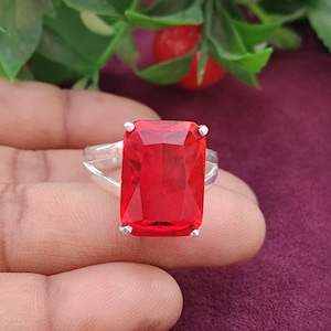 May include: A silver ring featuring a large, rectangular, faceted red gemstone set in a four-prong mounting. The band is split into two thin, polished silver segments that meet at the base of the stone, creating a simple and clean design.
