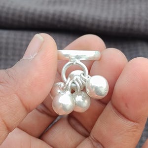 May include: A silver ring with a cluster of small, round, dangling charms. The ring and charms have a polished, reflective surface. The charms are spherical and hang from the ring's band.