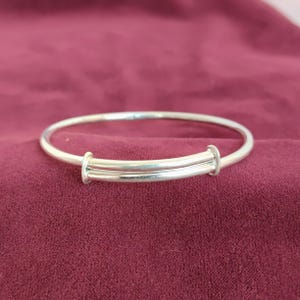 Peut inclure: Un bracelet jonc réglable argenté. Le bracelet a un design simple avec un mécanisme coulissant pour ajuster la taille. Le bracelet est présenté sur une surface bordeaux.