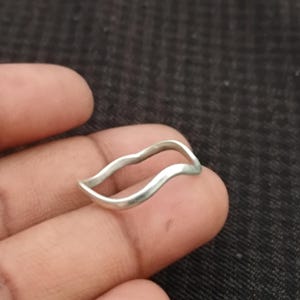 May include: A silver-colored ring with a wavy design. The ring has a smooth, polished finish and a simple, elegant style. The ring is held on a finger against a dark background.