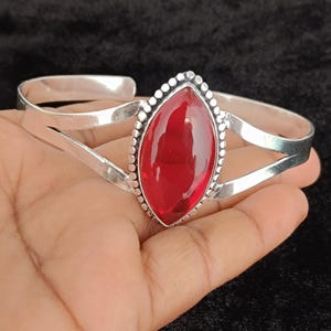 Pode incluir: Uma pulseira de prata com uma grande pedra vermelha translúcida em forma de marquise no centro. A pedra é emoldurada por uma borda de prata com contas, e a pulseira consiste em dois braços de prata polida divididos que se unem.