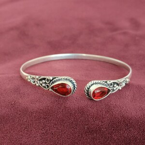 Peut inclure: Un bracelet manchette en argent avec des détails ornés et deux pierres précieuses rouges en forme de goutte. Le bracelet est ouvert et posé sur un fond bordeaux. Les pierres précieuses sont d'un rouge vif.