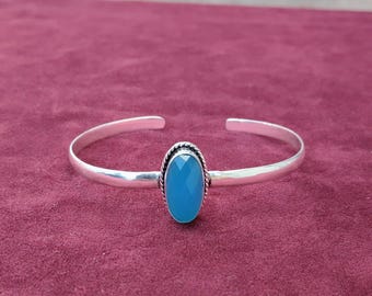 Könnte beinhalten: Ein silbernes Armreif-Armband mit einem großen, ovalen, facettierten, hellblauen Edelstein in der Mitte. Der Edelstein ist von einem gedrehten, seilartigen Design umgeben. Das Armband wird auf einer weinroten Oberfläche präsentiert.