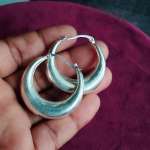 Op de afbeelding: Een paar gepolijste zilveren oorringen met een dik, afgerond profiel en een scharniersluiting. Het metalen oppervlak is glad en reflecterend, en aan de binnenzijde van een van de ringen staat duidelijk het 925-keurmerk voor sterling zilver.