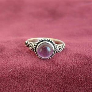 Może przedstawiać: Srebrny pierścionek z dużym, okrągłym, fioletowym kamieniem szlachetnym. Pierścionek ma ozdobną, sznurowatą obwódkę wokół kamienia szlachetnego i ozdobne wiry na obrączce. Pierścionek jest na bordowym tle.