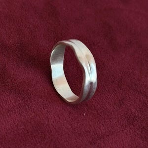 May include: A silver ring with a wavy design. The ring is made of a shiny, reflective metal and sits on a dark red surface. The ring's design is simple yet elegant, with a smooth, polished finish.