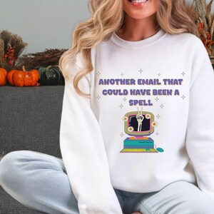 May include: A white long-sleeve sweatshirt featuring the purple text "ANOTHER EMAIL THAT COULD HAVE BEEN A SPELL" above a graphic of a vintage computer monitor displaying a skeleton hand making a peace sign, surrounded by small white flowers.