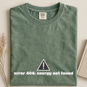 May include: A sage green crew neck t-shirt featuring a white graphic of a triangular warning sign with an exclamation point inside. Below the icon, the text reads "error 404: energy not found" in a bold, white, sans-serif font style.