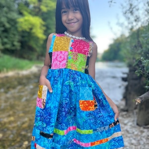 Puede incluir: Un colorido vestido patchwork con una falda azul y un corpiño con cuadrados amarillos, rosas y verdes. El vestido tiene bolsillos y se usa al aire libre cerca de un arroyo.