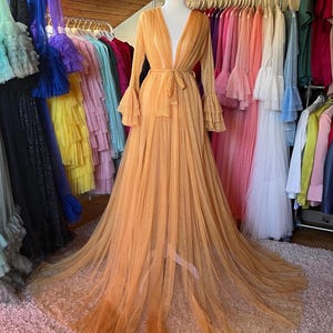 May include: An orange tulle gown with a plunging neckline and long sleeves with ruffled cuffs. The gown is tied at the waist with a matching sash and has a long, flowing train. A rack of colorful dresses hangs in the background.
