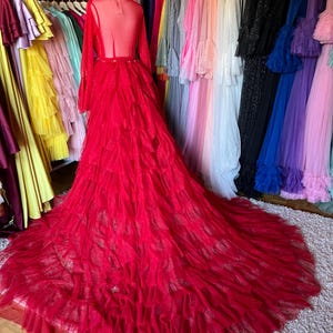Pode incluir: Um vibrante vestido de baile vermelho com uma longa cauda, exibido em um manequim. O vestido apresenta um corpete transparente, mangas compridas e uma saia com babados em camadas. Uma arara de vestidos coloridos está pendurada ao fundo.