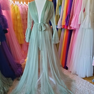 May include: A pale green tulle robe with long sleeves and a tie waist is displayed on a mannequin. The robe has a flowing, open front design. A variety of colourful tulle dresses hang in the background, creating a vibrant display of apparel.
