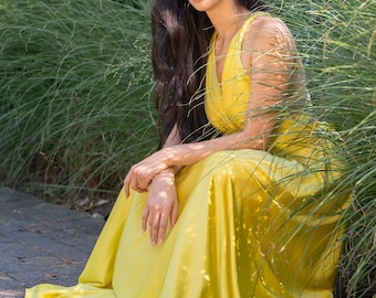 May include: A woman in a bright yellow maxi dress sits in a grassy area. She has long dark hair and is looking at the camera.