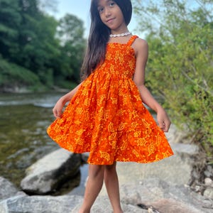 Puede incluir: Vestido de verano naranja con estampado de tortugas amarillas. El vestido tiene un corpiño smock y tirantes finos. La niña lleva un collar de conchas. El vestido llega a la rodilla.
