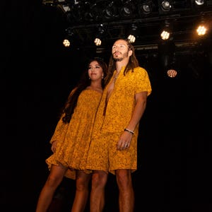 Puede incluir: Una pareja vistiendo atuendos a juego con estampado floral amarillo. La mujer lleva un vestido y el hombre lleva una camisa y pantalones cortos. Están de pie en un escenario con luces de fondo.