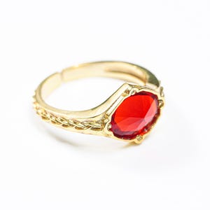 May include: A gold-toned ring featuring a central faceted red gemstone set within a decorative frame. The band displays a braided texture along the sides and includes an open-back design, allowing for adjustable sizing on the finger.
