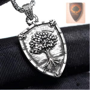 Peut inclure: Un pendentif en métal argenté en forme de bouclier, présentant un motif en relief d'un arbre avec des racines visibles. Le pendentif est suspendu à une chaîne en argent. Une petite image insérée montre un bouclier similaire sur fond orangé.
