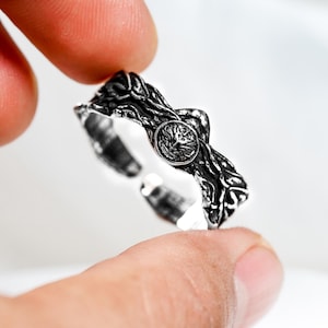 Puede incluir: Un anillo ajustable de tono plateado con un acabado oscuro oxidado y un diseño texturizado similar a ramas. La banda incluye un emblema circular central que representa un rostro felino estilizado, con un diseño abierto en la parte posterior.