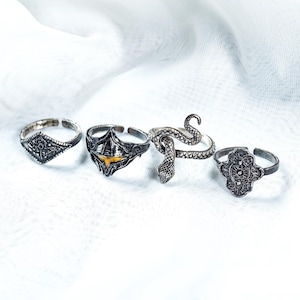 May include: A set of four silver-toned metal rings featuring distinct designs: a diamond-shaped engraved band, a helmet-shaped ring with a yellow accent, a coiled snake-shaped ring, and a quatrefoil-shaped ring with intricate embossed detailing.