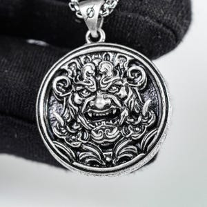 Può includere: Un ciondolo circolare color argento con un rilievo dettagliato di una creatura mitica ringhiante con zanne prominenti e motivi di capelli vorticosi. Il ciondolo è fissato a una catena metallica e presenta una finitura scura e ossidata.