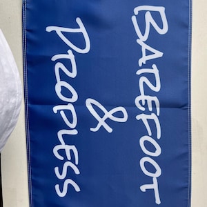 Pode incluir: Uma bandeira azul com texto branco "Barefoot & Progress". A bandeira tem uma borda branca na parte superior com dois ilh&oacute;s de metal. A bandeira est&aacute; pendurada em uma parede.