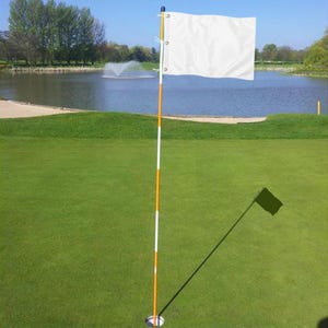 Pu&ograve; includere: Una bandiera da golf bianca tinta unita fissata a un'asta alta e sottile con strisce orizzontali alternate arancioni e bianche. L'asta &egrave; inserita in un foro circolare nell'erba verde, con la bandiera che si estende orizzontalmente a destra.