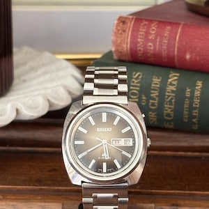 May include: A vintage Orient watch with a silver-tone case and band. The watch face is a gradient brown color with silver hands and markers. The date is displayed at the 3 o'clock position. The watch is displayed on a wooden stand.