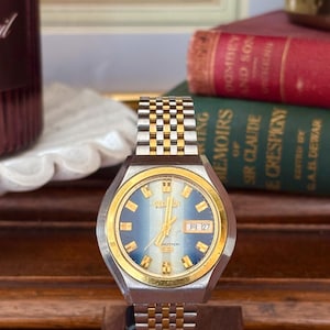 May include: A vintage Citizen watch with a gold and silver band and a blue face. The watch displays the date as "月27". The watch is set on a wooden stand, with books in the background.