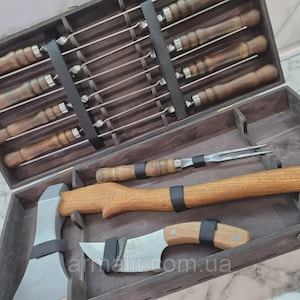 Puede incluir: Un juego de caja de madera oscura que contiene brochetas, un tenedor para trinchar, una hacha y un cuchillo. Las brochetas tienen mangos de madera y varillas de metal. El hacha y el cuchillo tienen mangos de madera y hojas de metal.