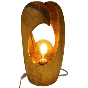 Puede incluir: Una lámpara de mesa de madera con un brillo cálido. La lámpara presenta un diseño único y orgánico con un marco de madera tallada. La bombilla es visible a través de una abertura ovalada. La lámpara está conectada a un cable de alimentación.