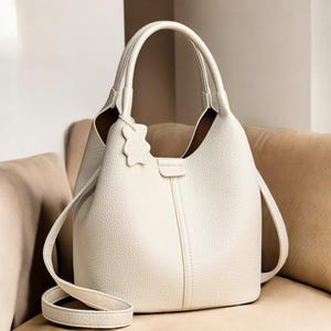 Könnte beinhalten: Cremefarbene Lederhandtasche mit einem oberen Griff und einem abnehmbaren Schulterriemen. Die Tasche hat eine strukturierte Oberfläche, einen kleinen Bärenanhänger und eine vertikale Naht auf der Vorderseite. Der Innenraum ist dunkler.