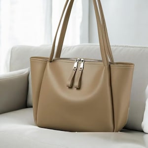 Puede incluir: Un bolso tote de color beige con dos asas largas y un cierre de cremallera plateado. El bolso tiene forma rectangular con esquinas redondeadas y una textura lisa similar al cuero. El bolso está sobre un sofá blanco.