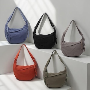 Peut inclure: Cinq sacs à bandoulière en forme de croissant, de couleurs variées : pervenche, noir, gris, orange et vert olive. Chaque sac est doté d'une longue bandoulière réglable et d'une fermeture éclair. Des détails tressés ornent les côtés.
