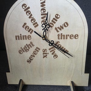 May include: A light-colored wooden clock with an arched top and two small support stands. The clock face features the words eleven, twelve, one, two, three, four, five, six, seven, eight, nine, and ten engraved in a circular pattern with black hands.