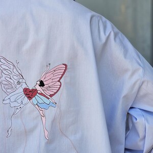 May include: Pale blue shirt featuring a butterfly fairy embroidery on the back. The fairy has pink and white wings, a blue skirt, and a red heart. The shirt has a blue collar and cuffs.