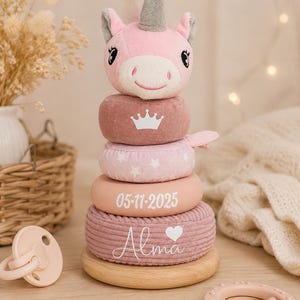 Può includere: Un giocattolo impilabile a forma di unicorno in peluche con testa rosa morbida e corno grigio. La pila presenta quattro anelli in tonalità rosa e malva, con una corona bianca, stelle bianche, la data 05-17-2025 e il nome Alma con un cuore.