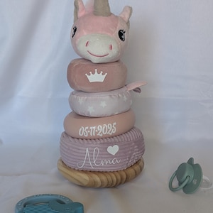 Puede incluir: Un juguete apilable de unicornio de peluche rosa y blanco con base de madera. El juguete tiene una cabeza de unicornio rosa, una corona, estrellas, la fecha "05-11-2025" y el nombre "Alma" impresos en los anillos. También hay un mordedor azul y un chupete verde.