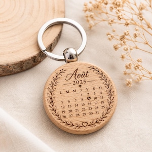 Peut inclure: Un porte-clés rond en bois gravé d'un calendrier pour le mois d'août 2025. Le motif présente une bordure de feuilles de laurier, le texte "Août 2025" et un petit cœur marquant le chiffre 9. Il est fixé à un anneau en métal argenté.