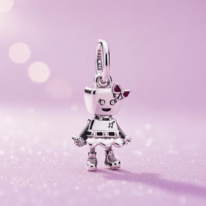 May include: A silver robot charm with a red bow and a white dress. The charm has a loop at the top for a necklace or bracelet. The charm is on a pink glittery surface.